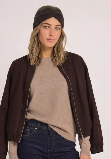Eine Frau trägt einen beigen Pullover, eine dunkelbraune Jacke, Jeans und ein schwarzes Stirnband.