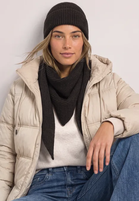 Junge Frau in beigefarbener Steppjacke, schwarzem Dreieckstuch und Mütze vor hellem Hintergrund.