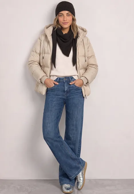 Frau in beiger Daunenjacke, schwarzer Mütze und Schal, weißem Pullover, blauen Jeans und silbernen Schuhen.