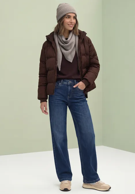 Beanie in Melange-Optik Muddy Mocha Frau in brauner Steppjacke, grauem Schal, Jeans und Mütze vor hellem Hintergrund.