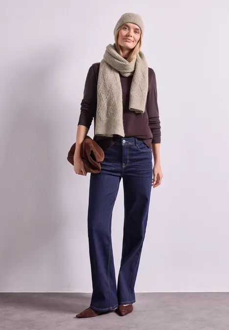 Frau trägt Wintermütze, Schal, Pullover und weite Jeans, hält Tasche, lässiger Look vor hellem Hintergrund.