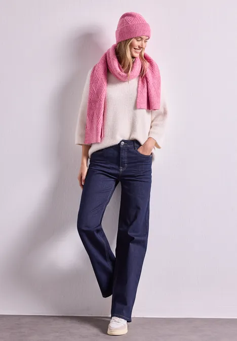 Frau in weißem Pullover, weiter Jeanshose, pinker Mütze und Schal steht vor hellem Hintergrund.