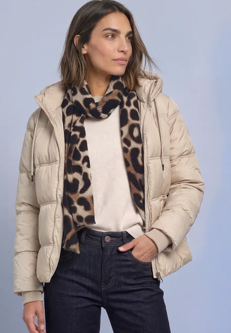 Vrouw in beige donzen jas, crèmekleurige trui, spijkerbroek en luipaardsjaal tegen effen achtergrond.