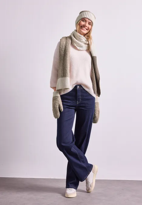 Frau in Winterkleidung mit Mütze, Schal, Handschuhen, hellem Pullover und Jeans vor neutralem Hintergrund.