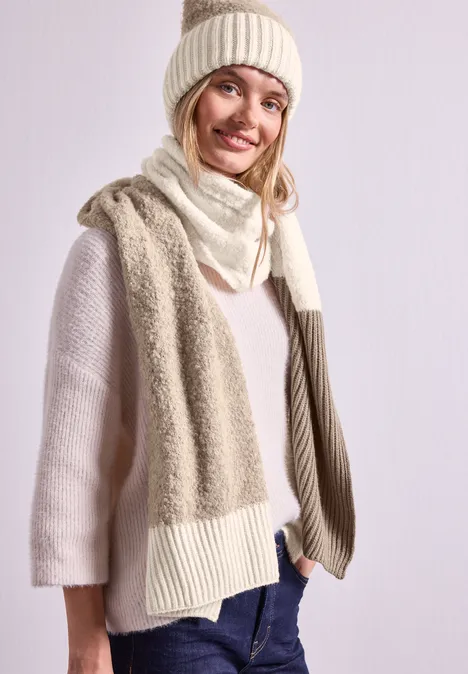 Eine Frau in cremefarbener Strickmütze, Schal und Pullover lächelt und trägt Jeans vor hellem Hintergrund.