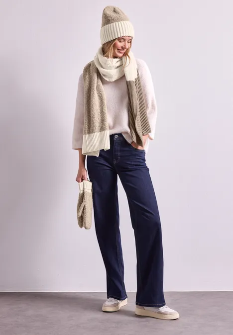 Frau in beiger Wintermütze, Schal und Strickpullover, weiten dunkelblauen Hosen, Hausschuhen und passender Tasche.