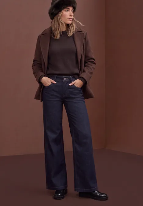 Frau mit braunem Blazer, Rolli, weiten Jeans und Hut vor braunem Hintergrund.
