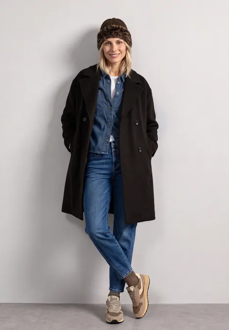 Muts met rand van imitatiebont in luipaardprint Black Vrouw met muts, overhemd, spijkerbroek, zwarte jas en sneakers staat ontspannen tegen een neutrale achtergrond.