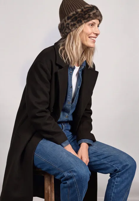 Muts met rand van imitatiebont in luipaardprint Black Vrouw zittend op kruk, zwart mantel, denim outfit en luipaardbont hoed, lacht opzij.