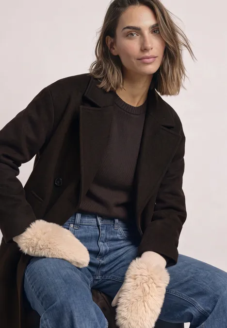 Vrouw zit ontspannen in een donkerbruine jas, bruin shirt, blauwe jeans en zachte wanten.