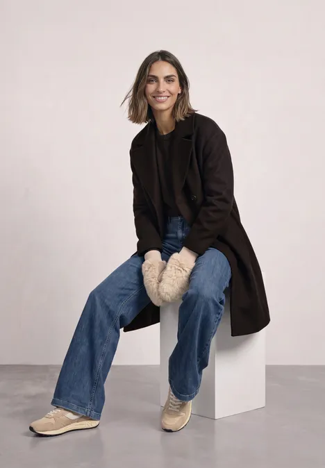 Een lachende vrouw zit op een wit blok, draagt een jas, jeans, gevoerde wanten en sneakers.