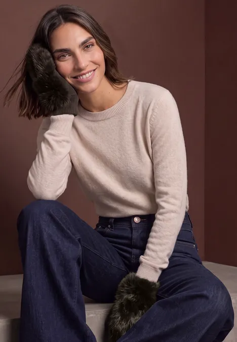 Jonge vrouw met donker haar in beige trui en spijkerbroek zit lachend voor een bruine achtergrond.