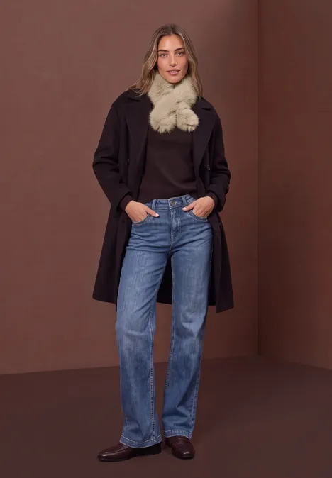 Effen stropdas van imitatiebont smooth stone sand Frau in dunklem Mantel, schwarzem Oberteil, Jeans und flauschigem Schal steht vor braunem Hintergrund.