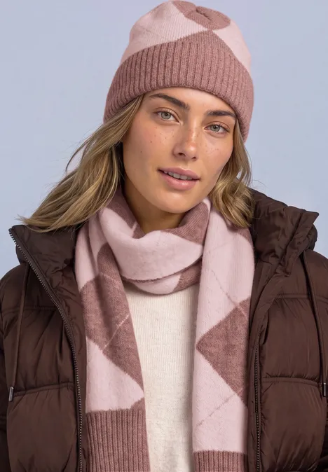Frau mit rosa-beiger Strickmütze und Schal trägt braune Steppjacke vor hellem Hintergrund.
