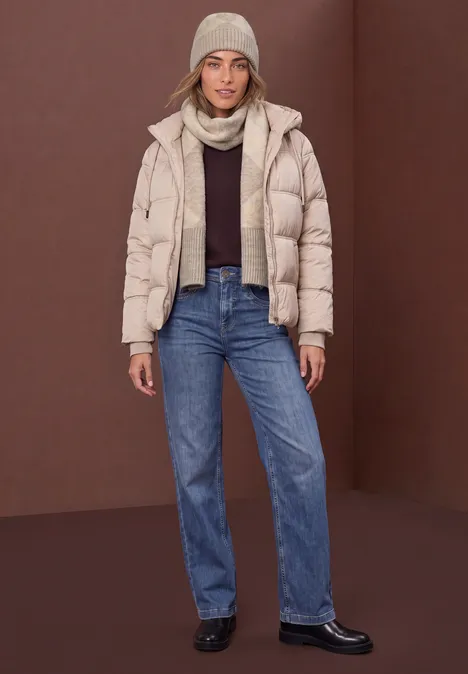 Frau in heller Steppjacke, Jeans, Mütze und Schal vor braunem Hintergrund.