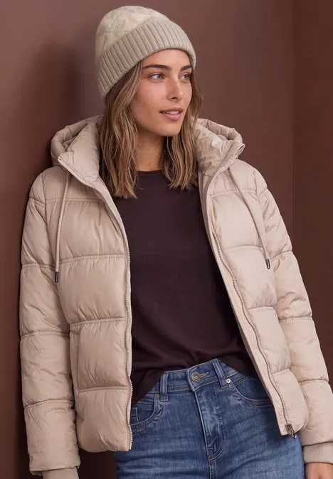 Frau in beiger Steppjacke mit Kapuze, Jeans und Strickmütze steht vor schlichtem Hintergrund.