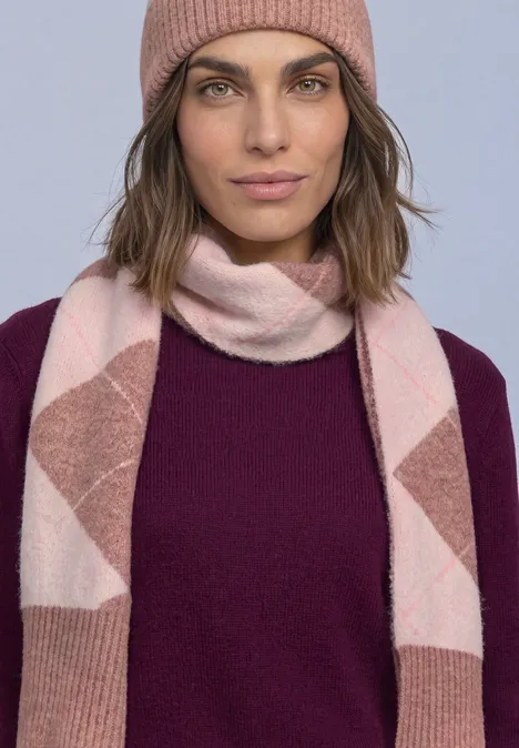 Longue écharpe à motif Argyle vintage blush mel. Frau mit rosa Beanie, rosa-weiß gemusterter Strickschal und dunkellila Pullover schaut ruhig in die Kamera.