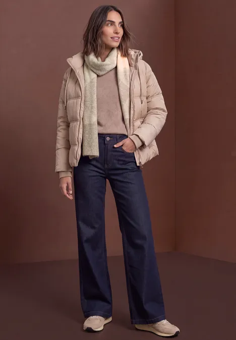 Longue écharpe à motif Argyle barley beige mel. Frau trägt beige Steppjacke, Schal, Pullover und weite dunkelblaue Jeans vor braunem Hintergrund.