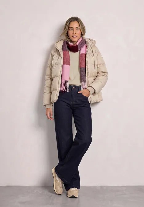 Frau trägt beige Steppjacke, Jeans, Schal und Sneakers in legerem Winterlook vor hellem Hintergrund.