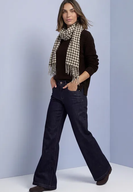 Écharpe à motif pied-de-poule et paillettes auburn brown Frau trägt braunen Pullover, Hahnentritt-Schal und weite, dunkle Jeans vor schlichtem Hintergrund.