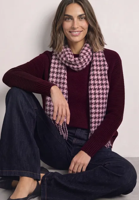 Écharpe à motif pied-de-poule et paillettes rustic rouge Frau in bordeauxrotem Pullover und karierter rosa Schal sitzt locker mit Jeans und lächelt.