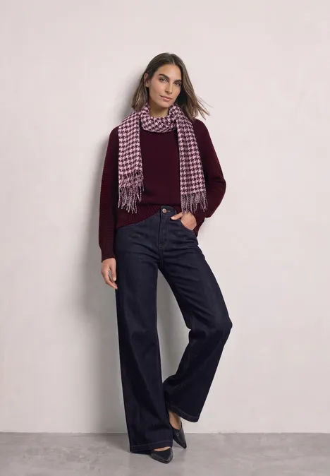 Écharpe à motif pied-de-poule et paillettes rustic rouge Frau in weinrotem Pullover, weitem Jeans, Hahnentritt-Schal und schwarzen Pumps vor hellem Hintergrund.