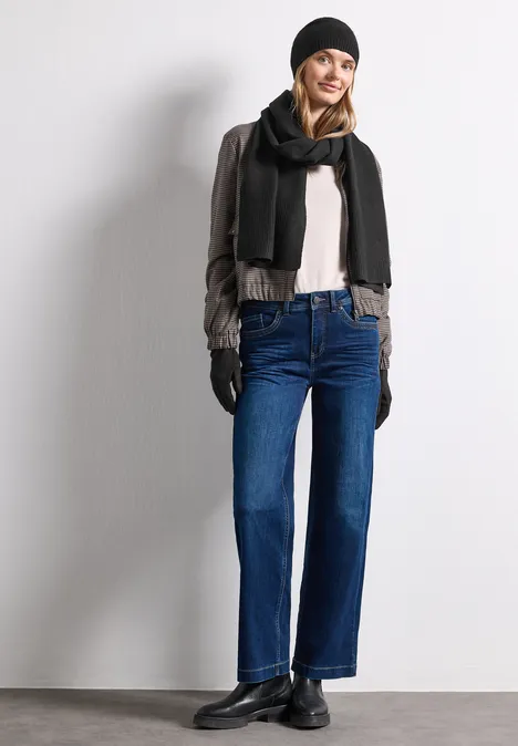 Feinstrick Handschuhe in Unifarbe Black Frau in Winteroutfit mit Mütze, Schal, Handschuhen, Jacke und geraden Jeans lehnt an Wand und lächelt.