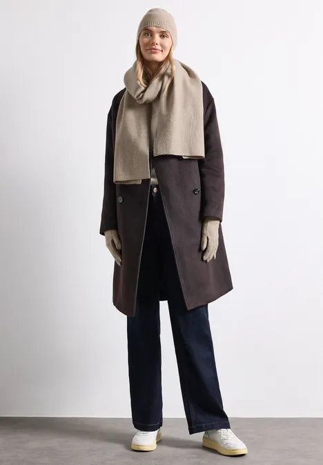 Eine Frau trägt Wintermantel, Schal, Mütze, Handschuhe und Hosen in neutralen Tönen.