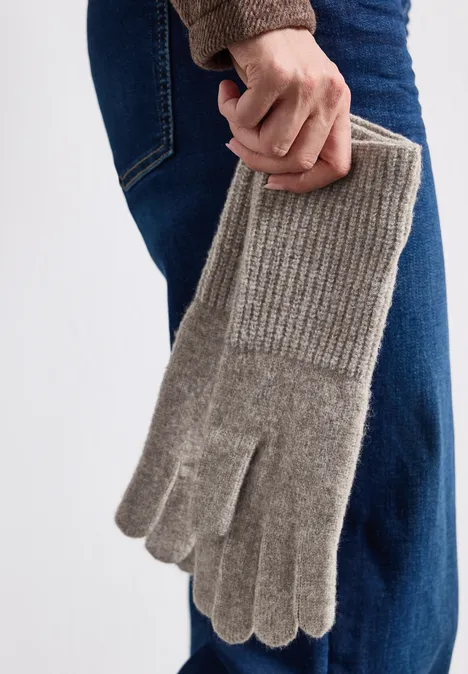Person hält graue, gestrickte Handschuhe mit Rippenbündchen, vor Jeans und Pullover. 