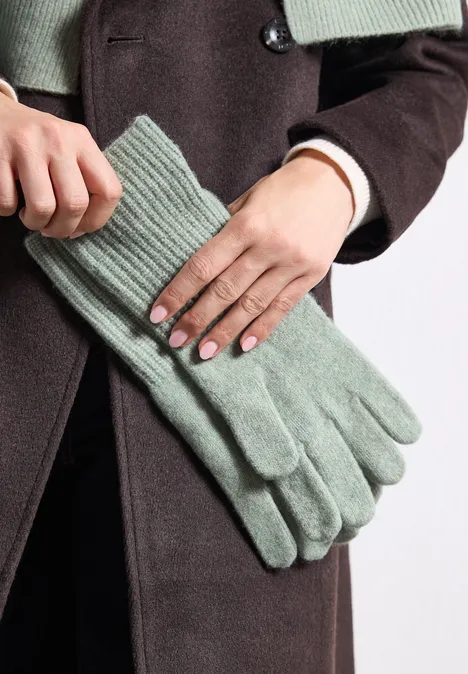 Eine Hand hält ein Paar hellgrüne Strickhandschuhe vor einem dunkelbraunen Mantel.