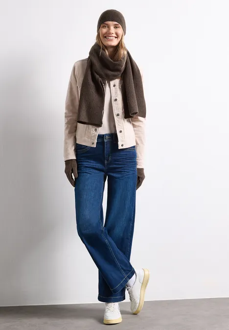 Eine Frau mit braunem Schal, Handschuhen, beiger Jacke und blauen Jeans steht vor weißem Hintergrund und lächelt.
