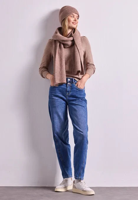 Junge Frau lehnt an weißer Wand, trägt Jeans, beigen Pullover, Schal, Mütze und Sneakers.