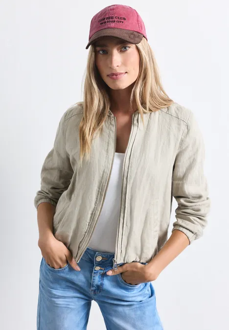 Junge Frau in heller Leinen-Blousonjacke, weißem Top, blauer Jeans und roter Kappe posiert lässig.