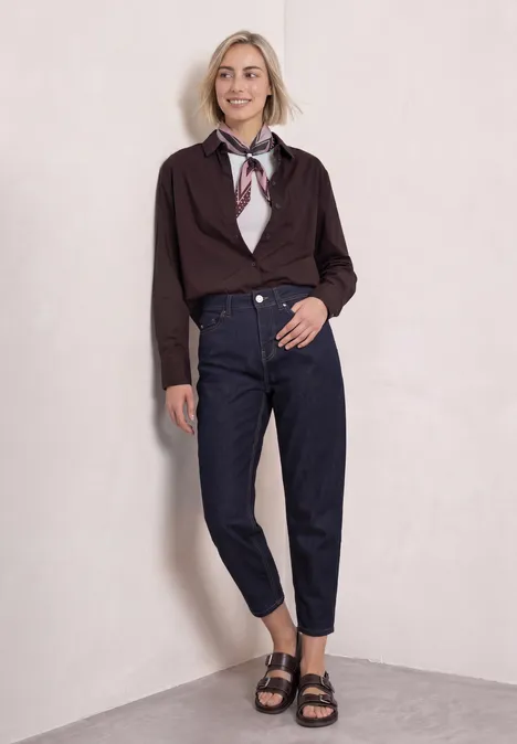 Junge Frau lehnt lächelnd an heller Wand, trägt dunkelblaue Jeans, Knotenbluse, Halstuch und schwarze Sandalen.