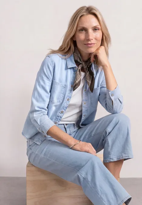 Eine Frau trägt eine helle Jeansjacke und Jeanshose, sitzt entspannt auf einem hölzernen Würfel.