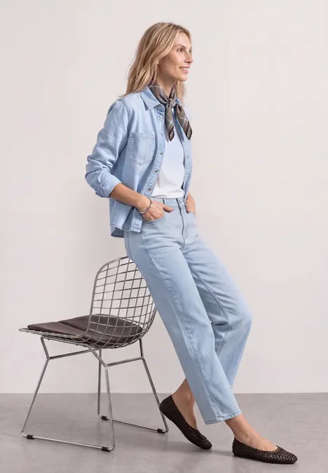 Eine junge Frau in hellem Jeansoutfit lehnt lässig an einem Drahtstuhl.