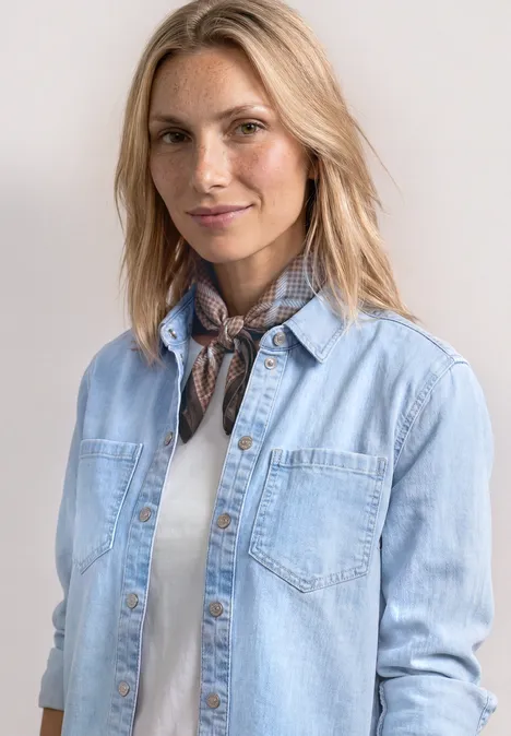 Frau mit hellblauer Jeansjacke, weißem T-Shirt und gebundenem Halstuch vor neutralem Hintergrund.