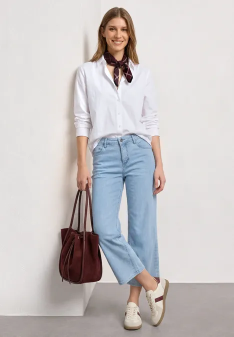 Eine Frau trägt weiße Bluse, blaue Jeans, weiße Schuhe und hält eine braune Tasche.
