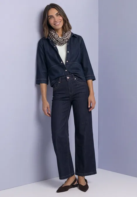 Frau lehnt an violetter Wand, trägt weite dunkelblaue Jeans, dunkles Hemd, Leoparden-Schal und flache Schuhe.