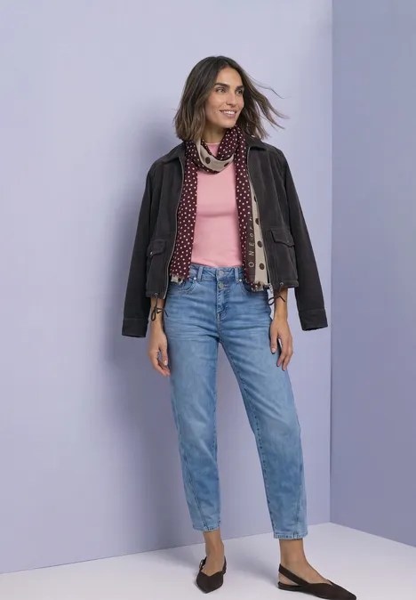 Frau trägt Lederjacke über rosa Oberteil, Schal, blaue Jeans und schwarze Schuhe vor lilafarbenem Hintergrund.