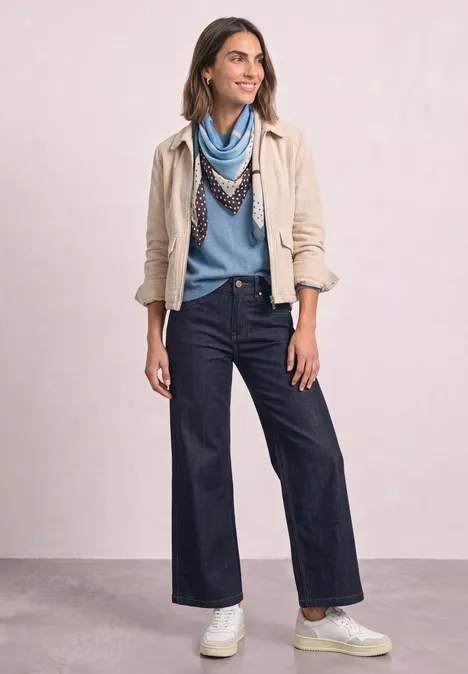 Frau in Denim-Culotte, hellblauem Pullover, hellbeiger Jacke, gemustertem Schal und weißen Sneakern.