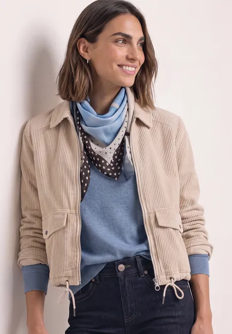 Frau in beiger Jacke, blauem Pullover und Tuch, lächelnd, vor hellem Hintergrund.