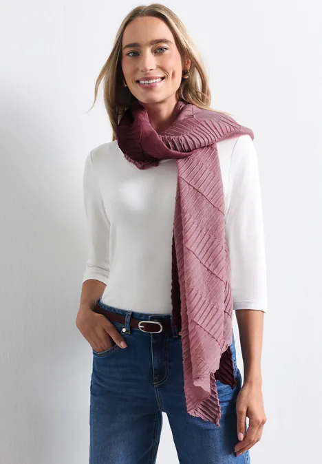 Lange sjaal met kleurverloop rustic rouge Eine lächelnde Frau in weißem Shirt, Jeans und weinrotem Strickschal vor hellem Hintergrund.
