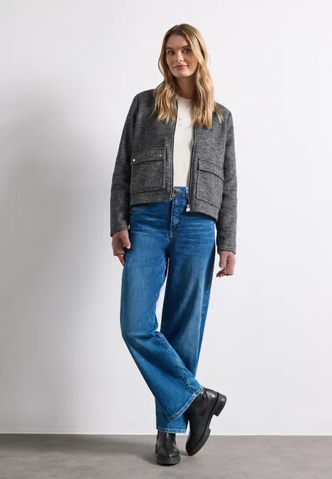 Junge Frau steht vor weißem Hintergrund, trägt graues Sakko, weißes Shirt und weite blaue Jeans.