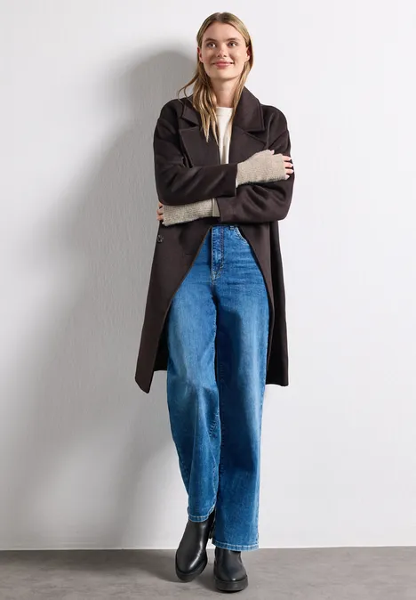 Eine Frau in schwarzem Mantel, weißem Shirt, blauen Jeans und schwarzen Boots steht vor weißer Wand.