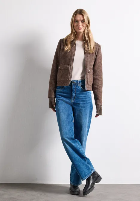 Eine Frau in brauner Jacke, beigem Shirt, blauen Jeans und schwarzen Stiefeln steht vor weißem Hintergrund.