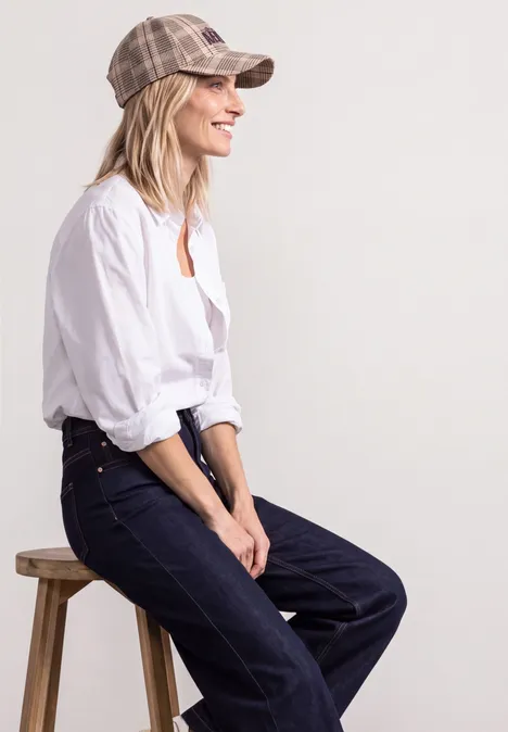 Pet met ruitjespatroon en slogan sanded amber Vrouw in wit overhemd, spijkerbroek en geruite pet, zittend op een kruk zijwaarts, glimlachend tegen een neutrale achtergrond.