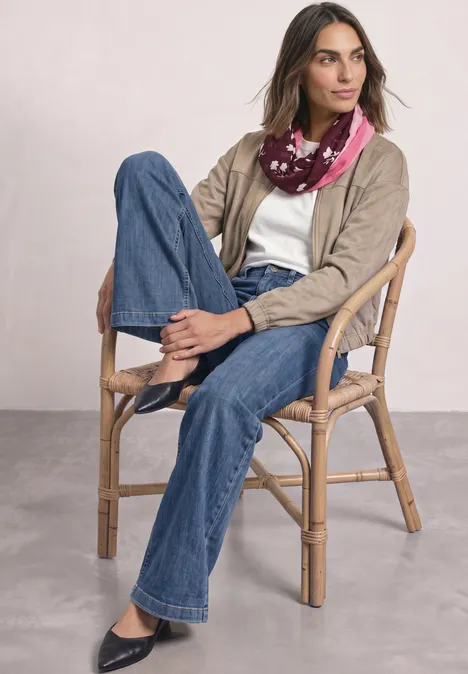 Loop-Schal mit floralem Muster rustic rouge Frau sitzt entspannt auf einem Korbsessel, trägt Jeans, T-Shirt, beige Jacke und bunten Schal.
