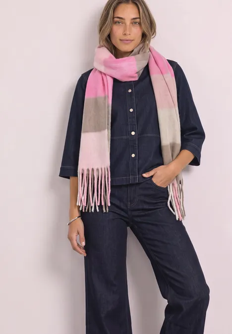Une femme porte une combinaison en jean bleu foncé et une écharpe rose et beige, debout sur fond clair.