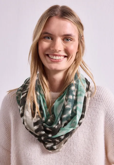 Eine junge Frau mit langen blonden Haaren lächelt, sie trägt einen grünen Schal und einen hellen Pullover.